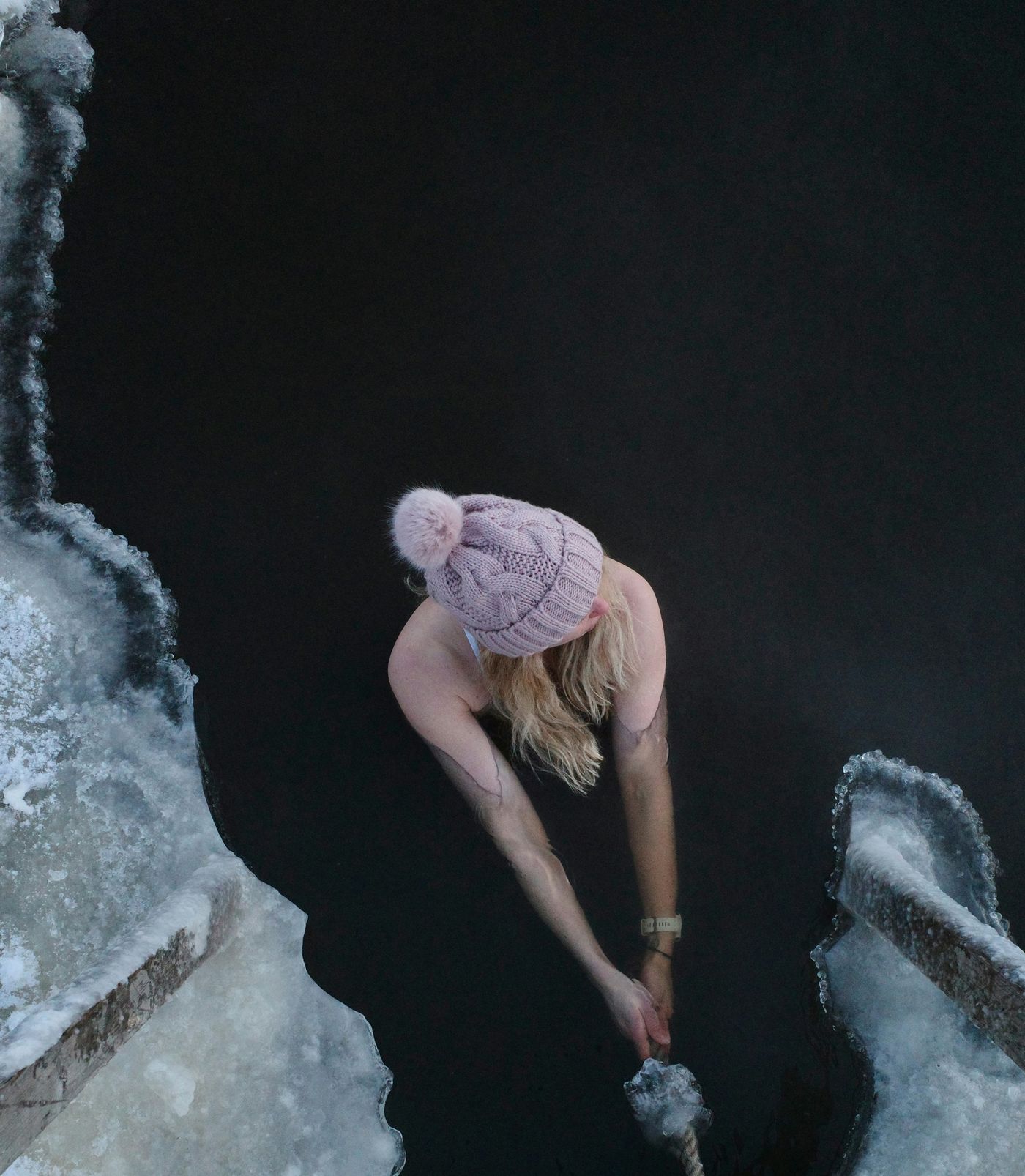 An overhead shot of a person wearing a pink knit hat taking a cold plunge in a hole cut into a frozen body of water.