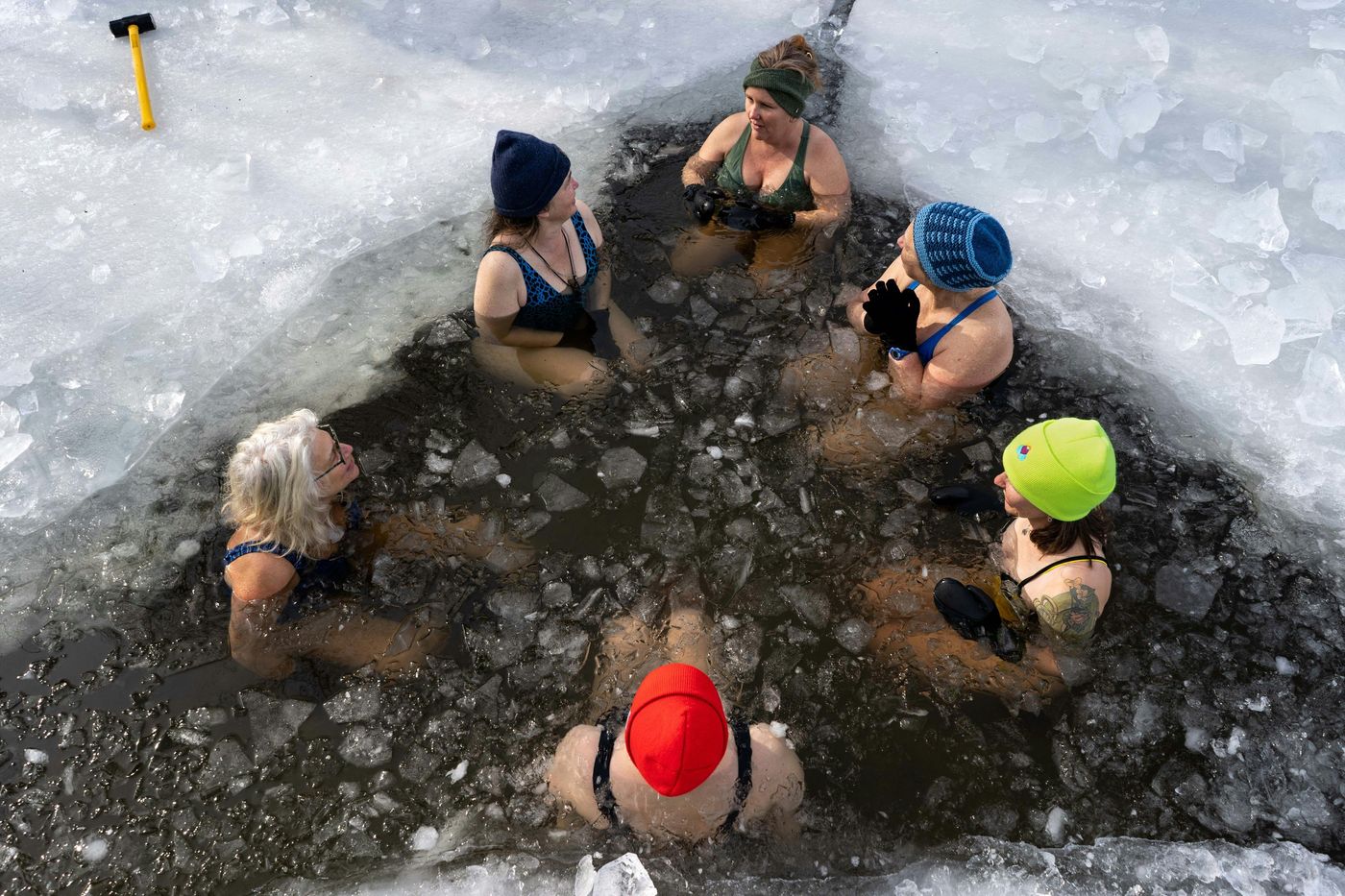 A group of people in swimsuits and winter hats are taking a cold plunge in a hole cut into a frozen lake or pond.