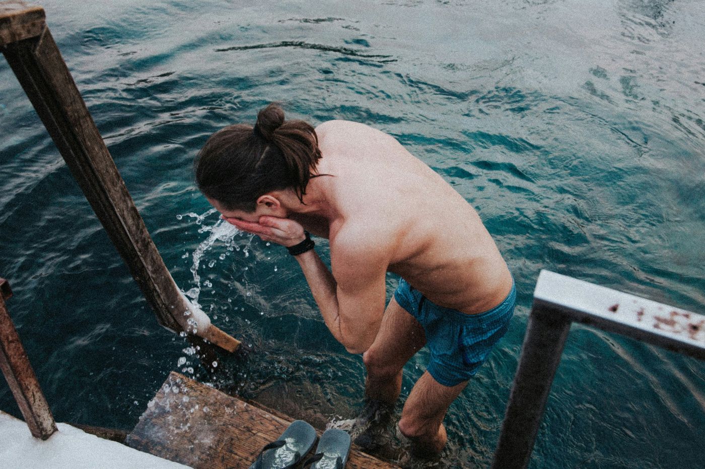 A person in swim trunks stands on wooden steps in a cold body of water, splashing water on their face.
