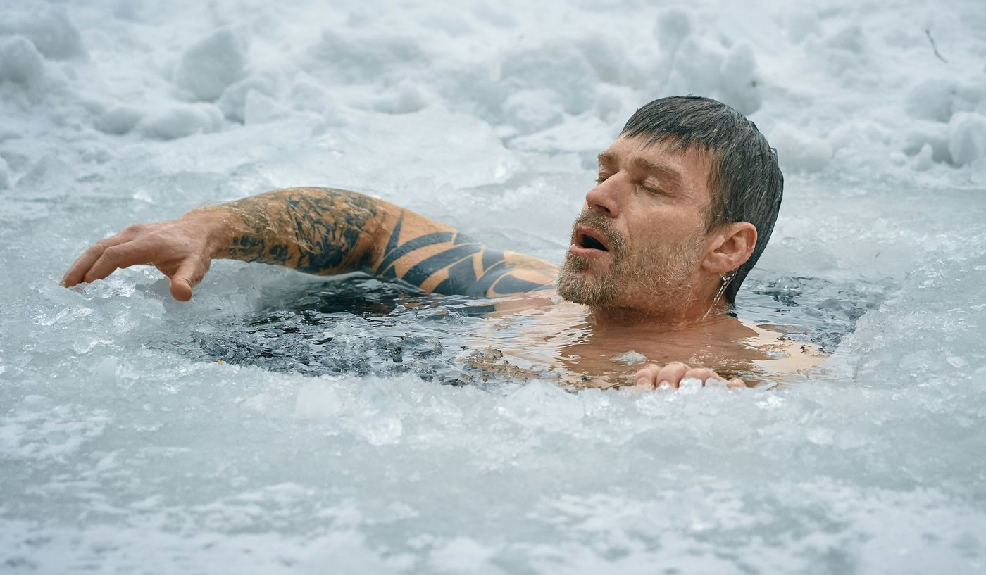 A man with a tattooed arm is submerged in an icy body of water, reacting to the cold.