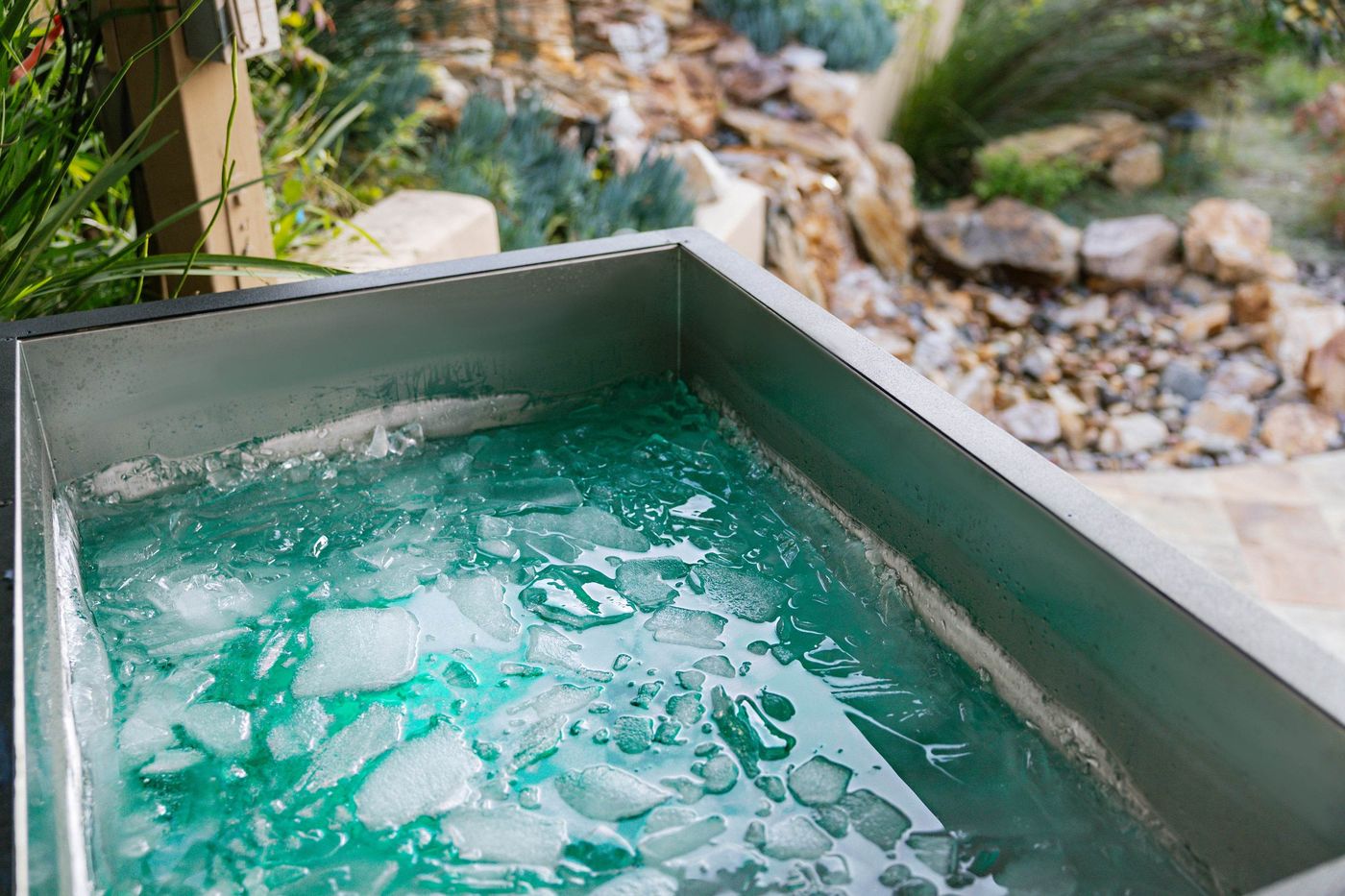 A close-up of a metal cold plunge tub filled with turquoise water and ice, set outdoors.