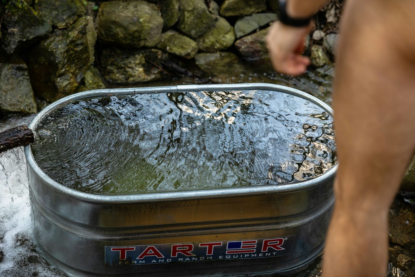A person next to a galvanized metal stock tank being filled with water in a rustic, rocky outdoor setting.