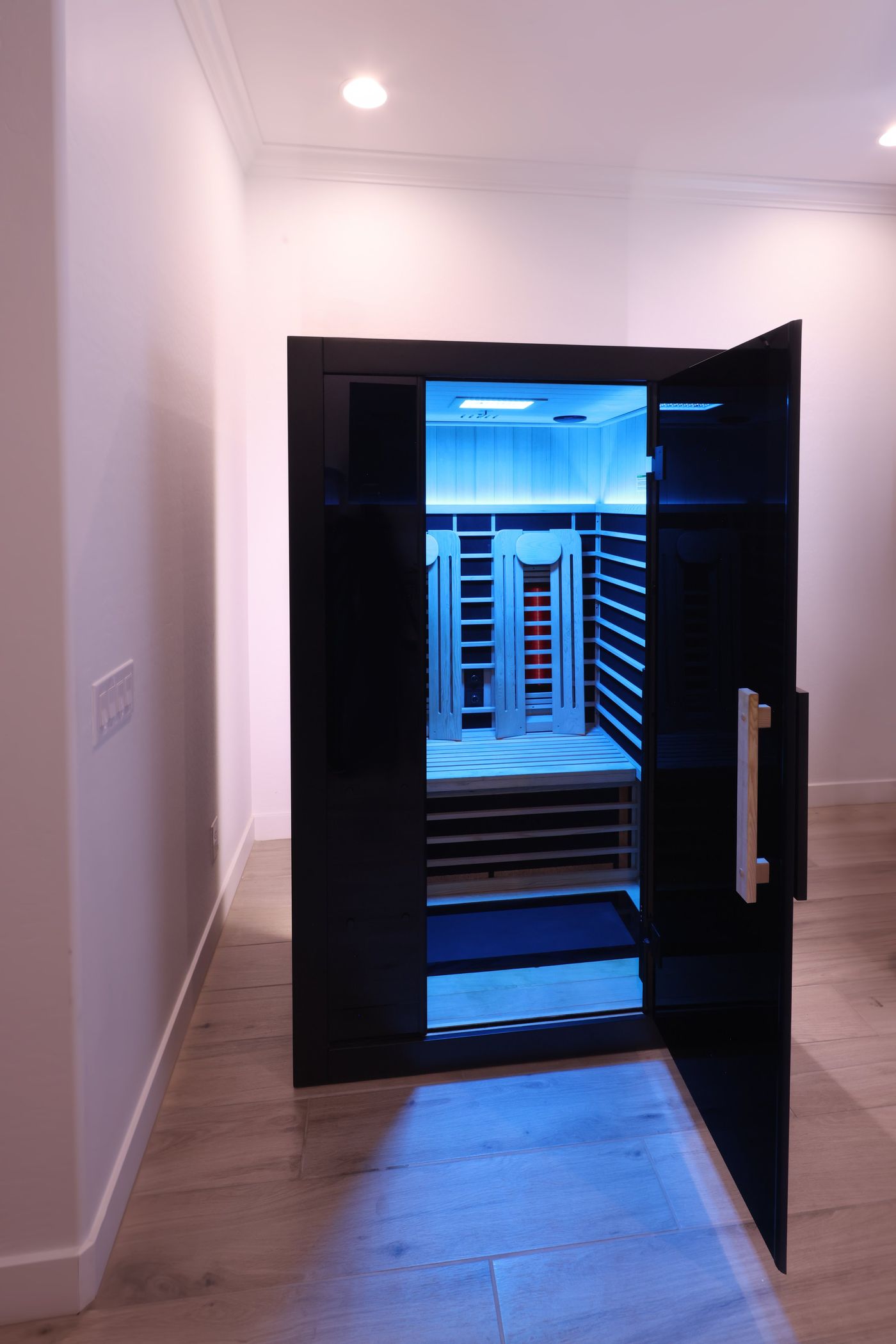 An empty, modern infrared sauna with its door open, showing a wooden interior with benches and blue lighting.