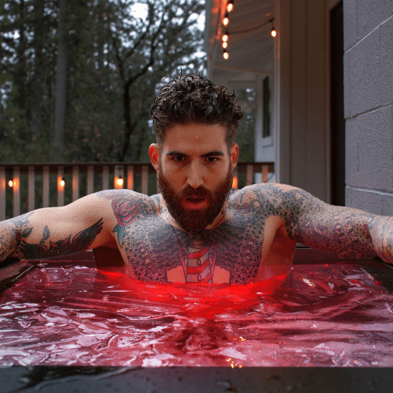 A tattooed man with a beard sits chest-deep in an outdoor cold plunge tub with an intense expression.